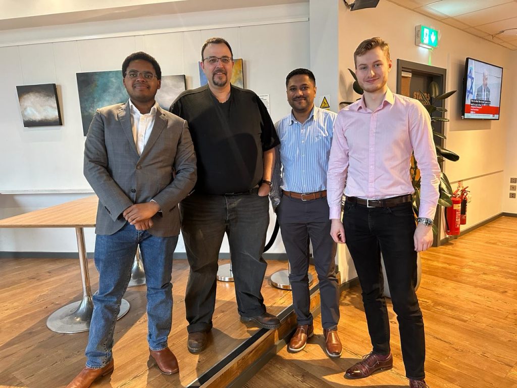 "Four men standing side by side, smiling, in an indoor setting with paintings in the background and a digital display screen to the right.